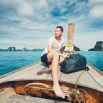 Young man (tourist) with backpack on the traditional long tail boat against islands between Phuket and Krabi in Thailand.